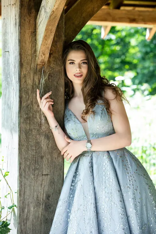 Woman in a pale blue gown standing beneath a wooden gazebo in a garden
