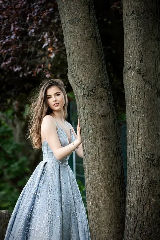 Woman in a pale blue gown standing beside a tree trunk