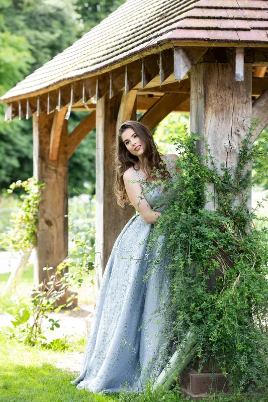 Woman in a pale blue gown walking through a garden past a wooden summerhouse