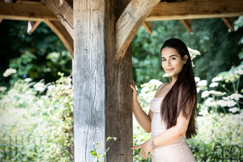 Woman in a pale gown standing beneath a wooden gazebo surrounded by greenery