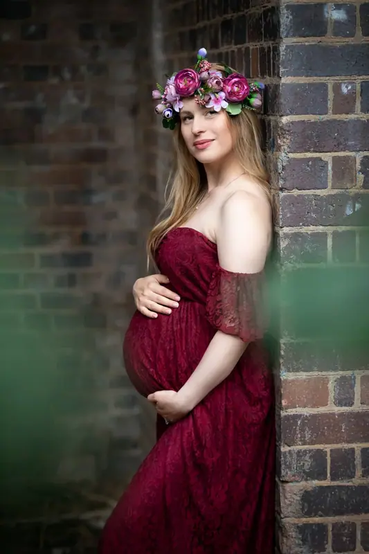 Expectant mother in a burgundy gown and flower crown beside a brick wall