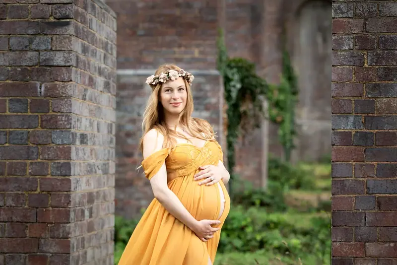 Expectant mother in a yellow gown and flower crown in front of an old brick building