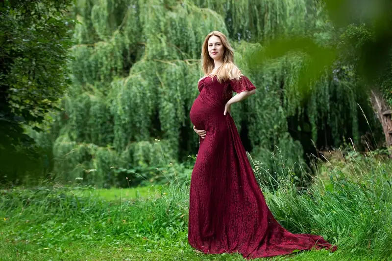 Expectant mother in a long burgundy gown standing beneath a willow tree