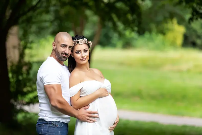 Expectant couple in white embracing beneath trees in a park
