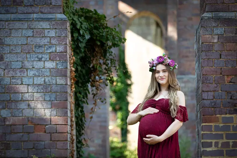 Expectant mother in a burgundy gown beside an ivy-covered brick wall