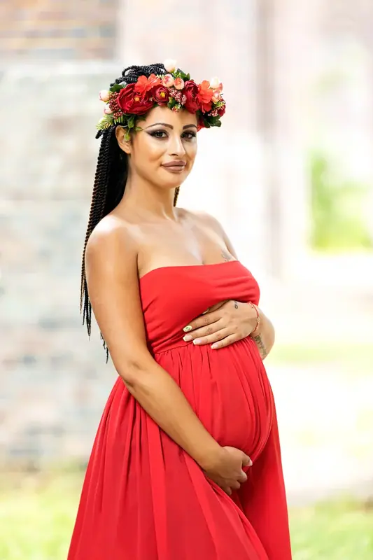 Expectant mother in a red dress and flower crown beside a brick wall