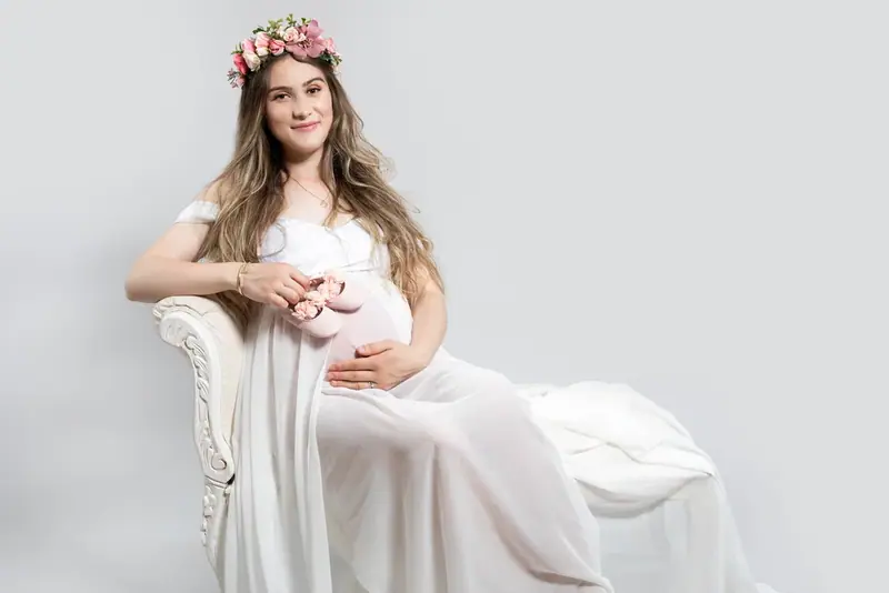 Expectant mother in a white gown and flower crown seated on a white armchair