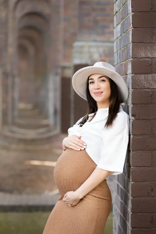 Expectant mother in a hat and fitted dress leaning against a brick pillar