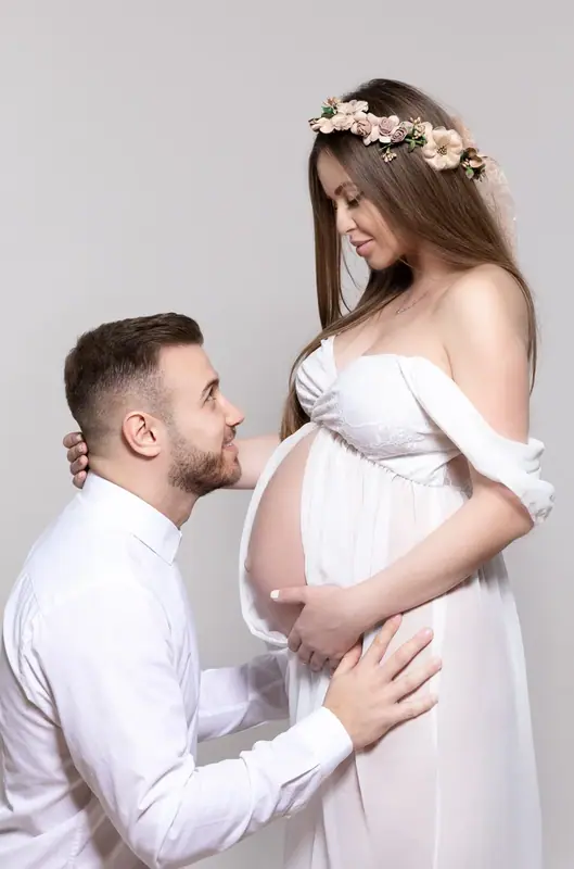 Expectant couple in white, the father with a hand on the bump, studio portrait
