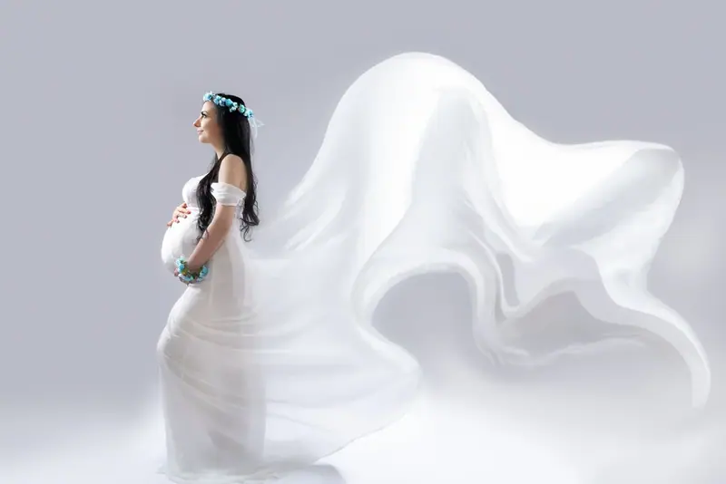 Expectant mother in a white gown with fabric billowing around her, white studio