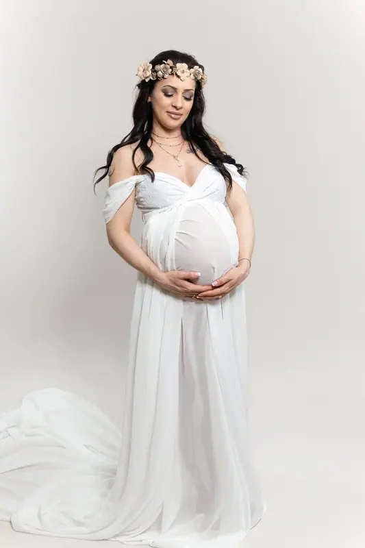 Expectant mother in a white off-the-shoulder gown and flower crown, white studio