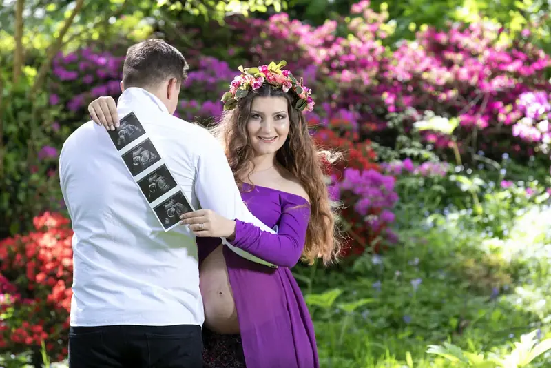 Expectant couple in purple and white holding ultrasound pictures among flowering shrubs