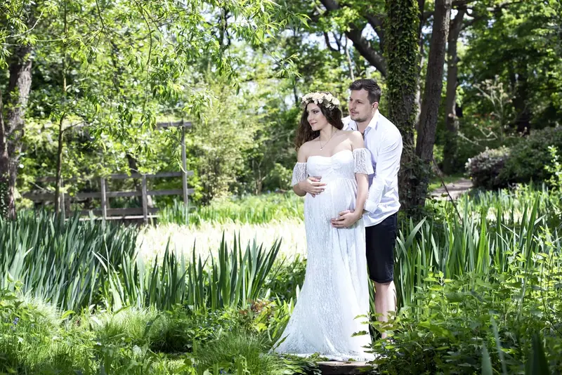 Expectant couple in white standing in a garden with a pond