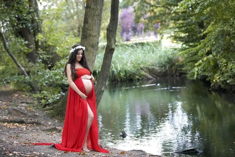 Expectant mother in a long red gown standing beside a garden pond