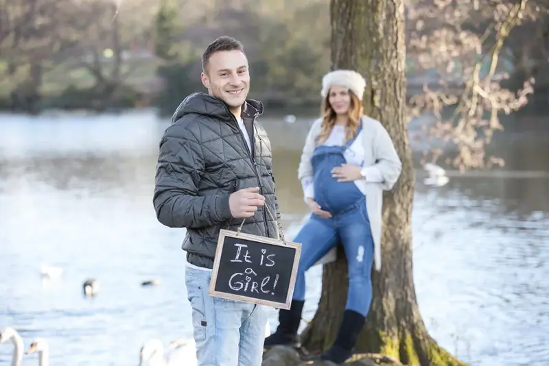 Expectant couple in winter coats holding a chalkboard sign beside a lake