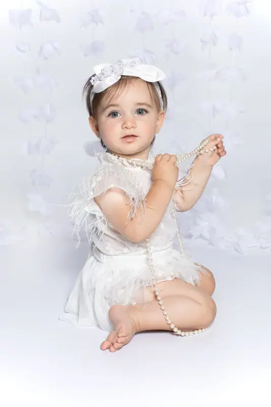 Baby girl in a white dress and flower headband sitting on a white backdrop