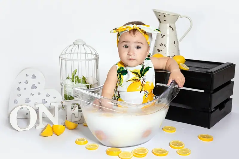 Baby sitting in a bowl of milk with lemon slices and a white jug, beside the word ONE