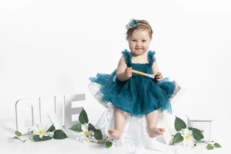 Baby girl in a teal dress sitting on a white backdrop beside wooden letters spelling ONE