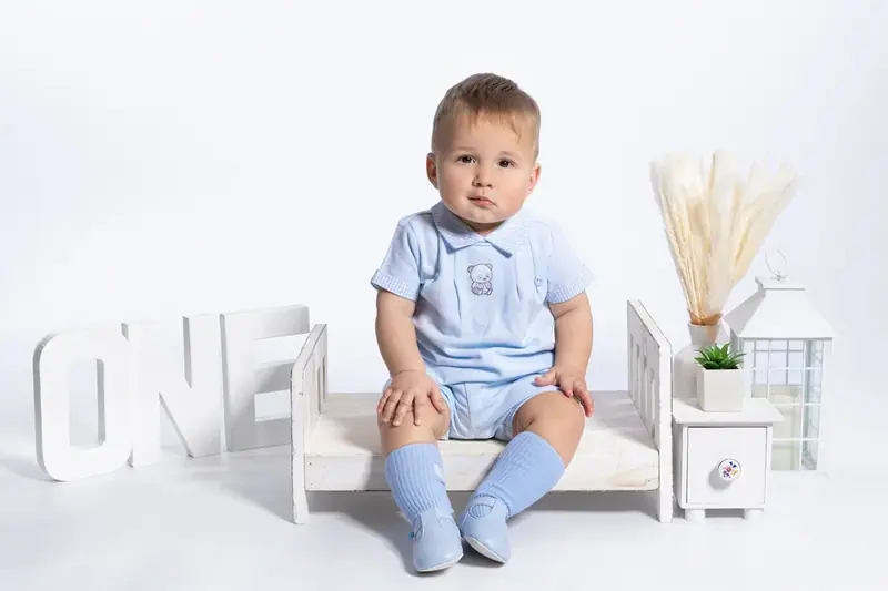 Baby boy in a blue outfit sitting on a small white bench beside the word ONE