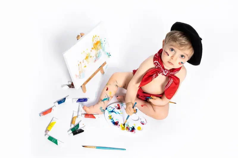 Baby in a beret and red neckerchief painting at a small easel with paint on the floor