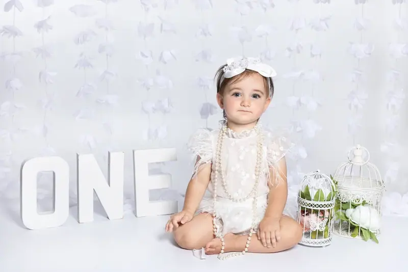 Baby in white sitting beside large letters spelling ONE on a pale backdrop