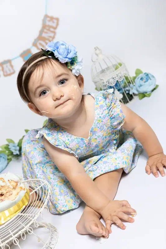 Baby in a blue floral outfit and headband sitting beside a birthday cake