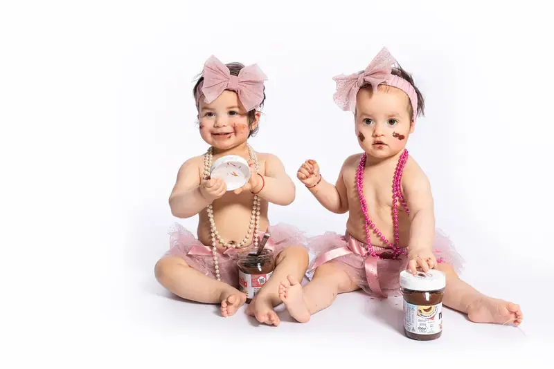 Two babies in bows and pearls sitting with small cakes on a white backdrop