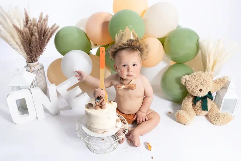 Baby with a cake surrounded by green and cream balloons and a teddy bear
