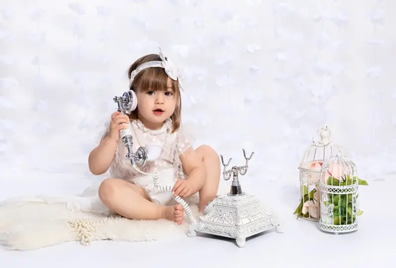 Baby girl in a white outfit sitting on a fur rug beside a lantern and a jug
