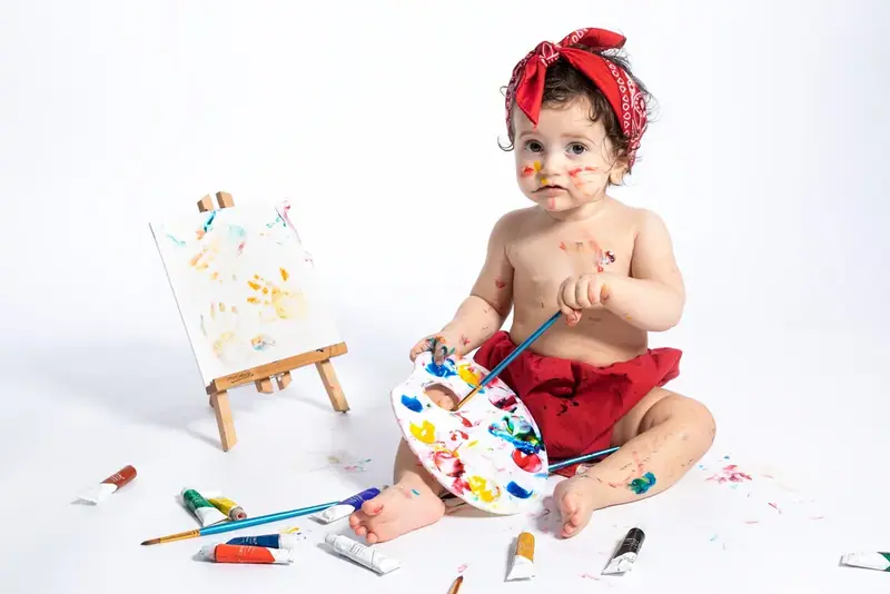 Baby with a red headband painting at a small easel with paint on the floor
