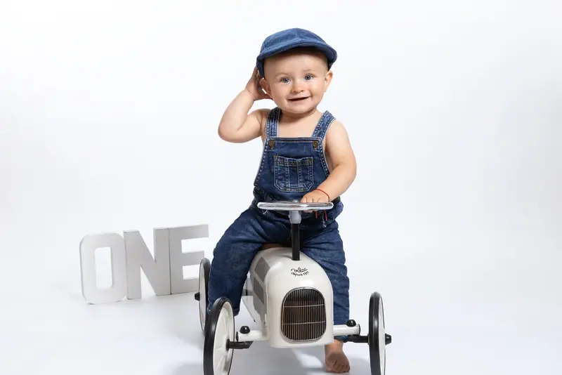 Baby in denim dungarees and a cap sitting on a toy car beside the word ONE
