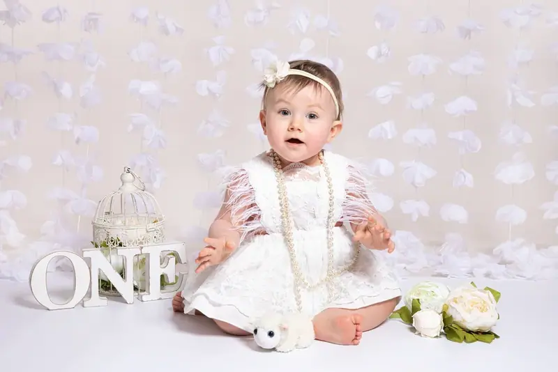 Baby in a white dress and headband sitting beside the word ONE on a patterned backdrop