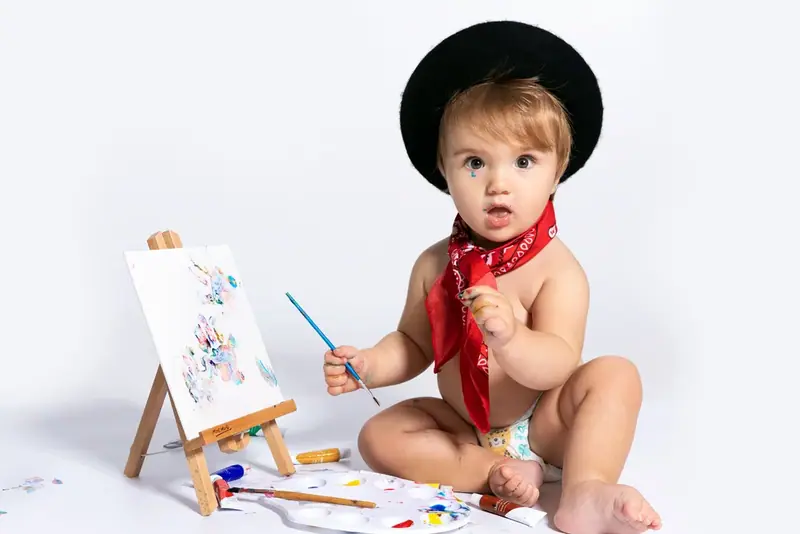 Baby in a black hat and red neckerchief painting at a small easel