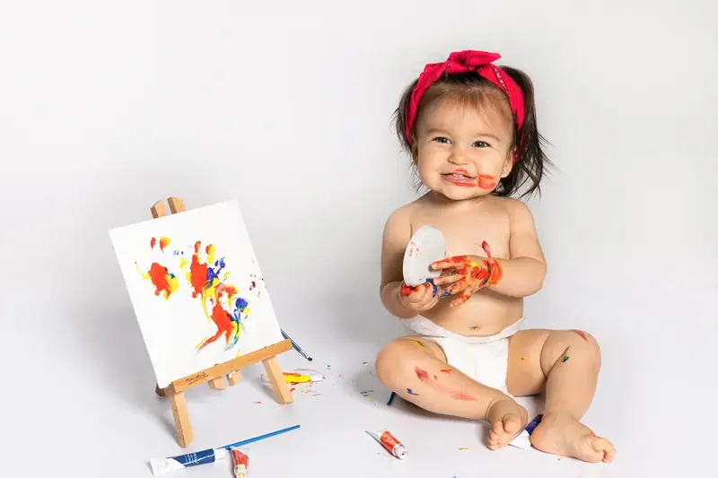 Baby with a red headband sitting beside a small easel with paint splashes