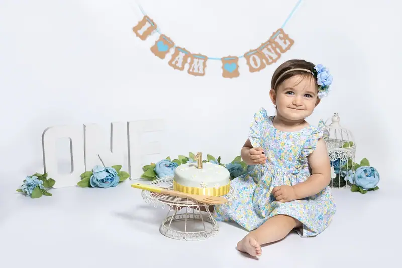 Baby in a blue floral dress sitting beside a cake and the word ONE under a bunting banner