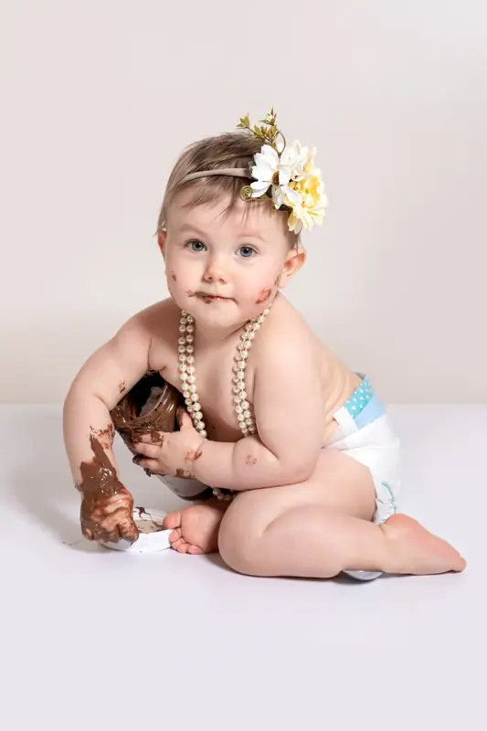 Baby in a nappy and a flower headband with pearls, holding a toy