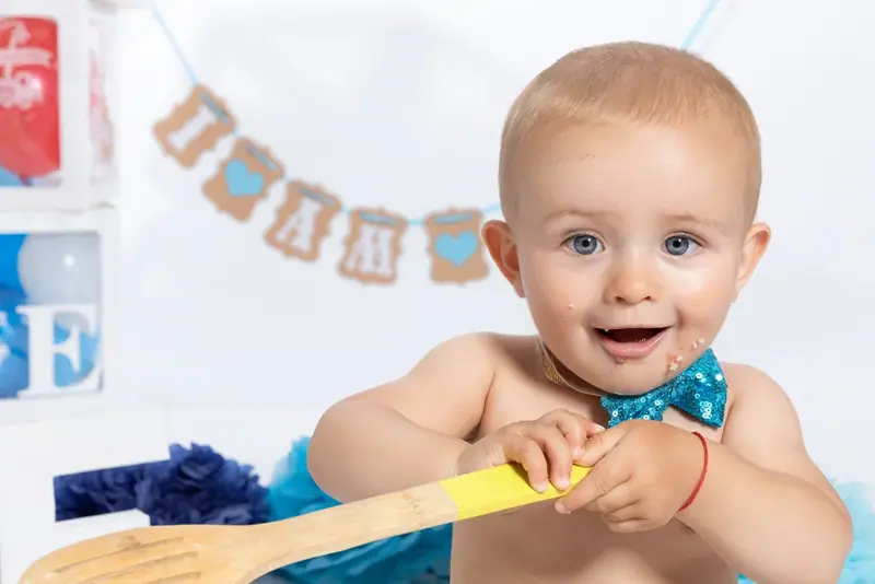 Smiling baby with a blue bow tie holding a wooden spoon beneath a bunting banner