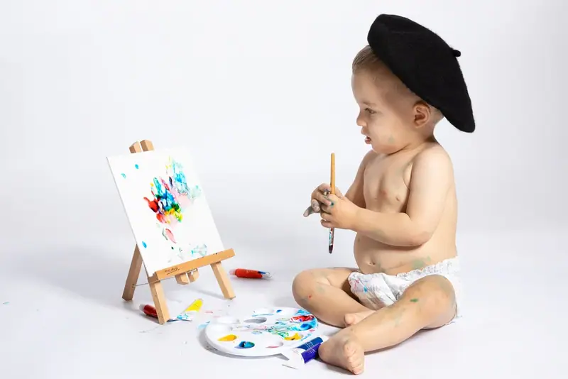 Baby in a black beret painting at a small easel on a white backdrop