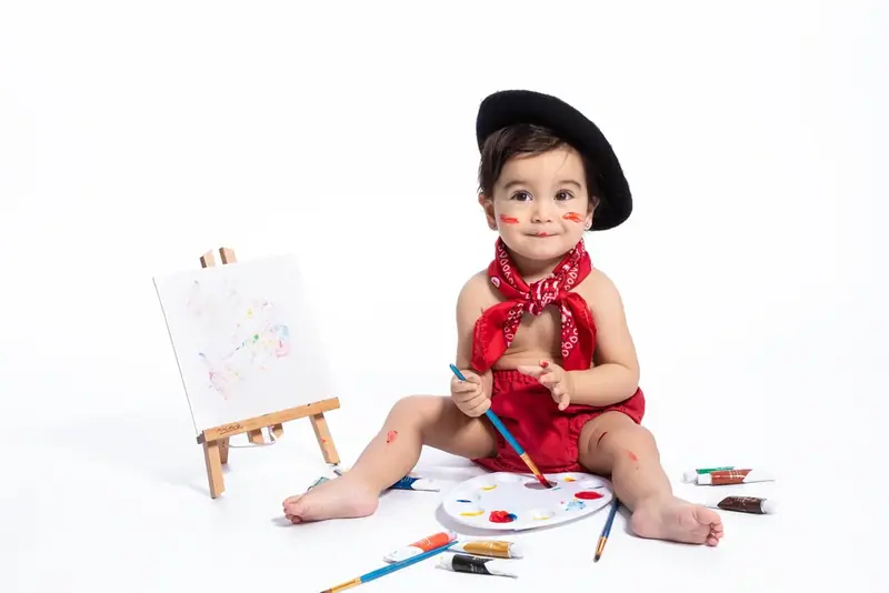Baby in a black hat and red neckerchief sitting beside a small easel