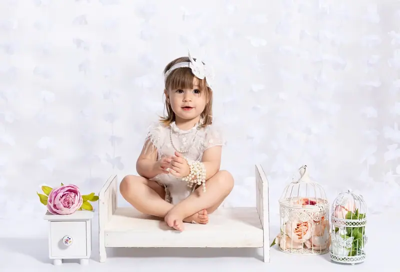 Baby in a white dress sitting on a small bench beside the word ONE