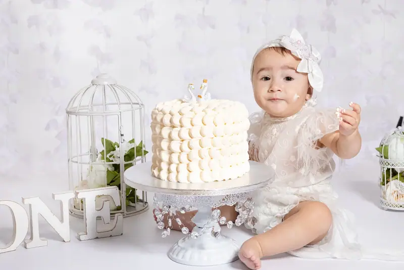 Baby in white sitting beside a white cake on a stand