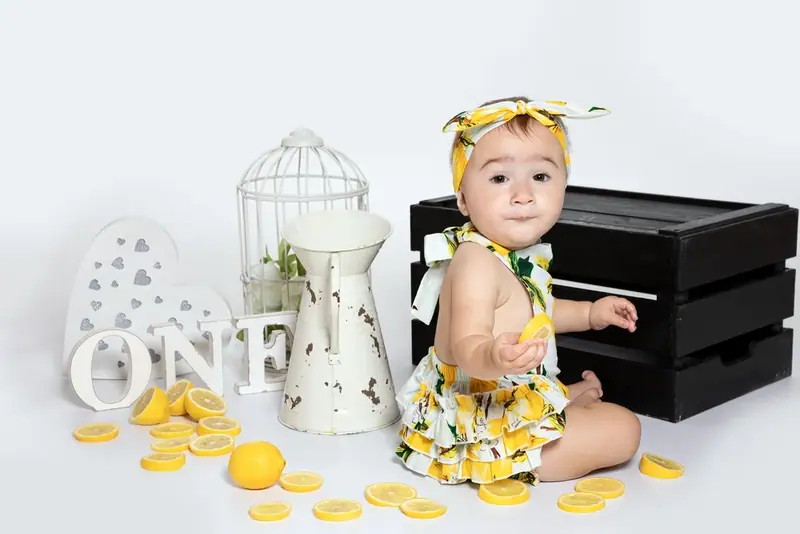Baby in a lemon-print outfit sitting beside a black crate, lemon slices and the word ONE