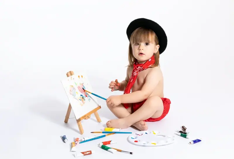 Baby in a black hat and red neckerchief sitting by a small easel with paint on the floor