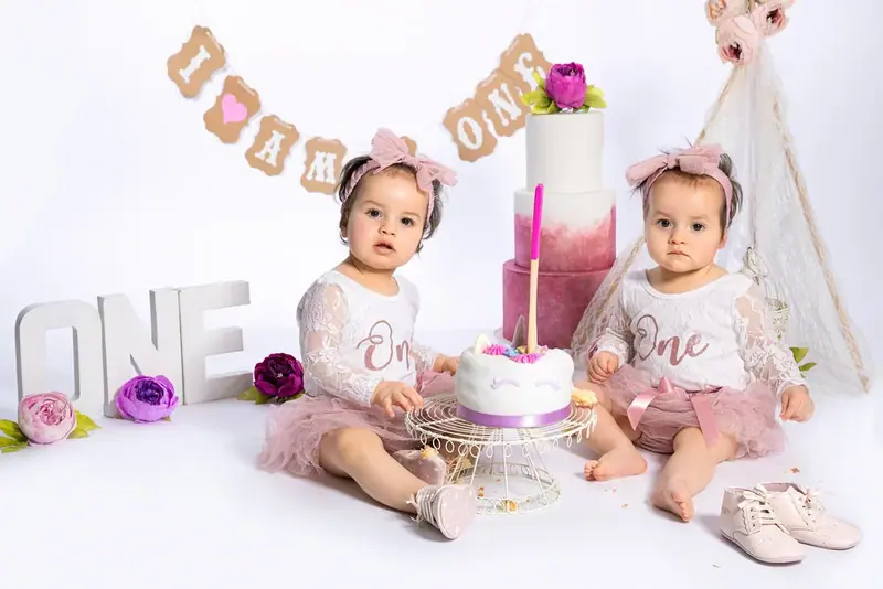 Baby in white beside a pink birthday cake, balloons and the word ONE