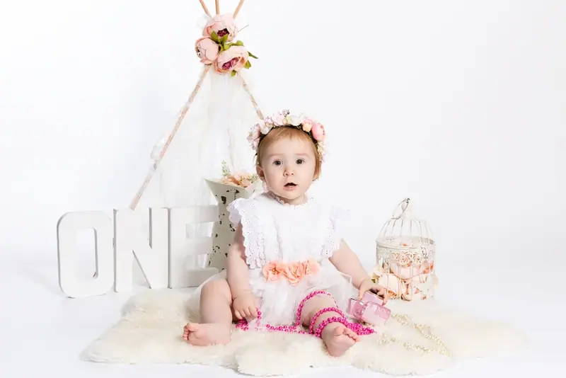 Baby in a white dress sitting under a teepee beside the word ONE