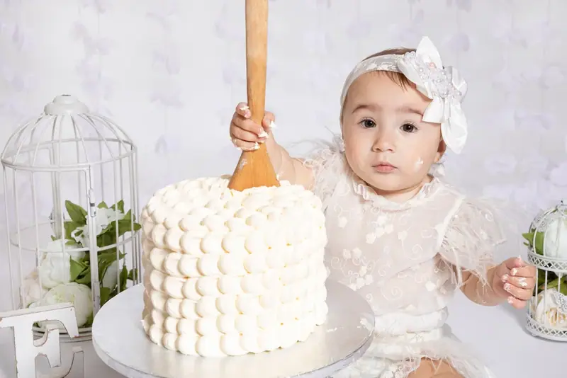 Baby holding a wooden spoon beside a white birthday cake