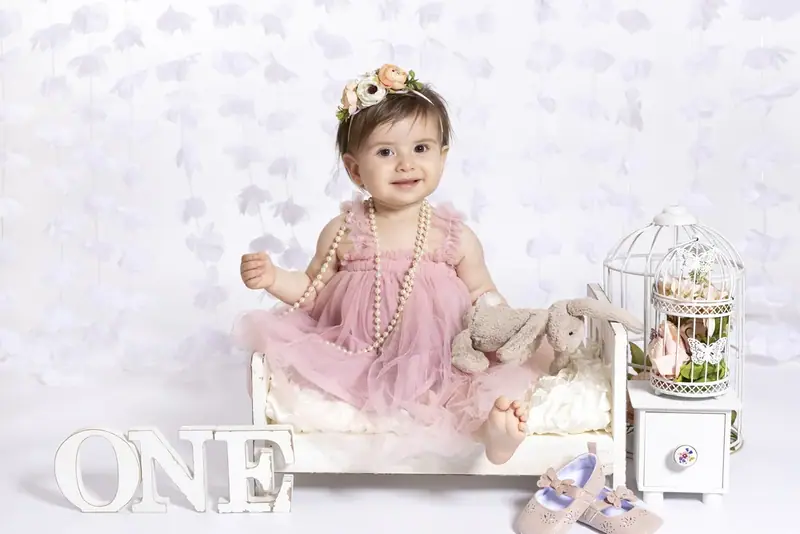 Baby in a pink dress sitting on a small bench beside the word ONE