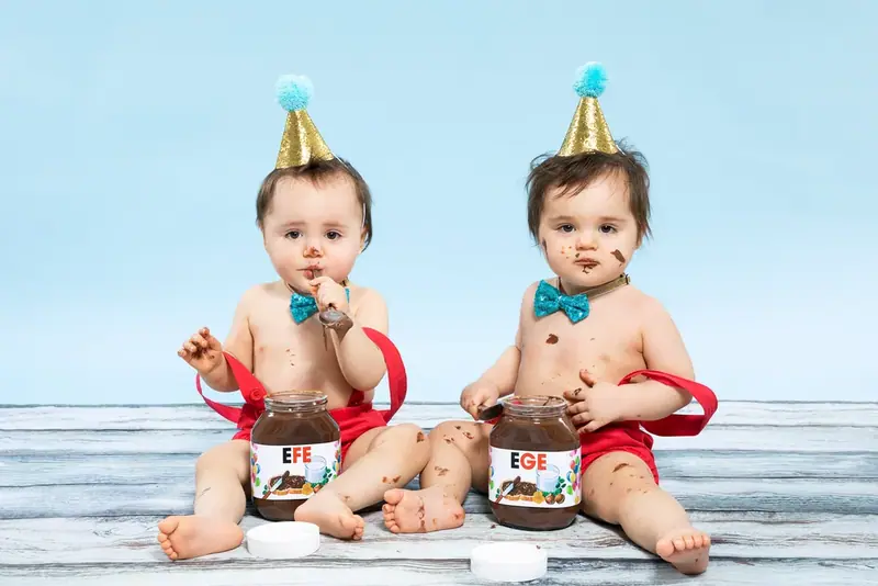 Two babies in party hats and bow ties sitting on a blue backdrop with cakes