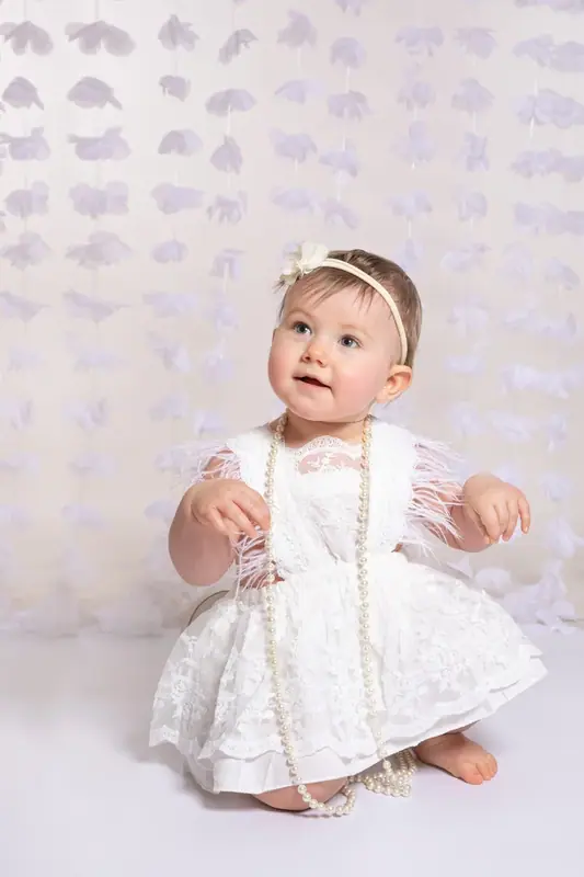 Baby in a white dress and headband sitting on a patterned backdrop