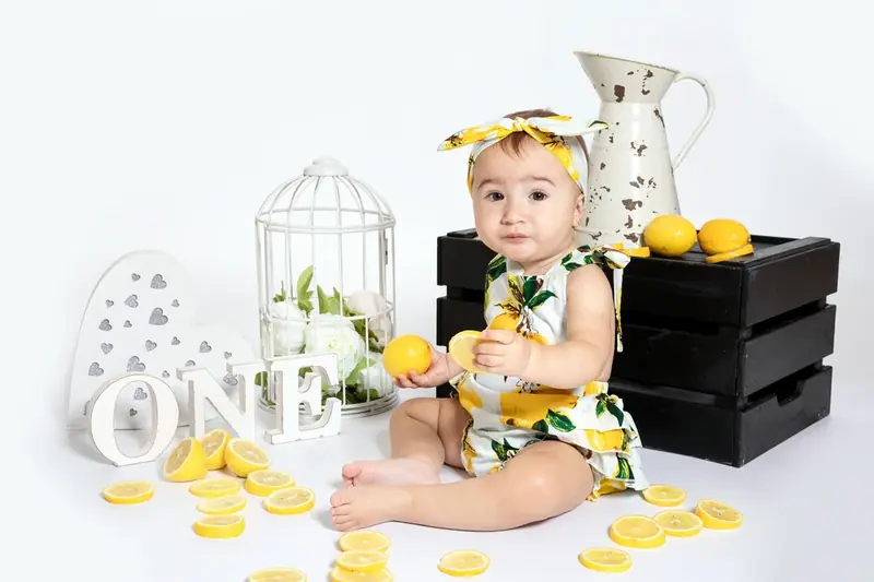 Baby in a lemon-print outfit sitting beside a black crate, jug and lemon slices